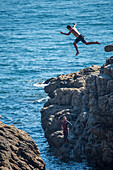 A young individual jumps off a rocky cliff into the sea in Ouistreham, Brittany, France, conveying a sense of adventure and excitement.