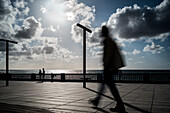 Stimmungsvolle Szene mit Silhouetten, die den Paseo de Santa Barbara in Cádiz, Andalusien, Spanien, entlanggehen. Die Sonne scheint durch dramatische Wolken über dem Meer.