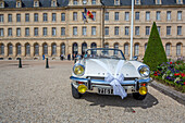 Vintage Triumph wedding car adorned with white ribbon, parked in front of Caen City Hall in Normandy, France. Elegant and classic wedding scene.