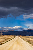 Empty dirt road leading to foothills under stormy skies