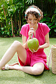 Young woman in pink short jumpsuit drinking coconut water from a coconut