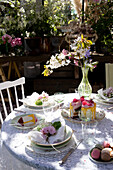 Frühlingshaft gedeckter Gartentisch mit Blumen und Gebäck auf weißer Spitzentischdecke