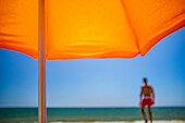 A relaxing summer scene featuring a bright orange umbrella with a blurred background of a man by the ocean.