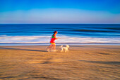 Artistic long exposure capturing a girl running with her dog along a beach at sunset. Movement and vibrant colors evoke joy and freedom.