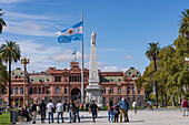 Menschen um eine argentinische Flagge & Pirámide de May oder Mai-Pyramide auf der Plaza de Mayo vor der Casa Rosada. Buenos Aires, Argentinien. Die Statue stellt die Freiheit dar