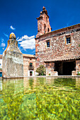 A tranquil drinking trough stands beside a historic building in La Barbolla, with clear water reflecting the vibrant blue sky and surroundings.