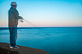 A blurred figure of a fisherman casting a line at sunrise in Ayamonte, Spain. The serene sea and sky create a peaceful and tranquil atmosphere.