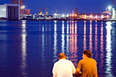 Two men enjoy the tranquil evening by the sea at the Colón promenade, overlooking the harbor in Barcelona.