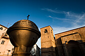 Ein kugelförmiger Springbrunnen steht auf der Plaza de San Nicolás mit der Kirche dahinter unter einem klaren blauen Himmel in Plasencia