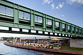 People relax on sunbeds along the Spree river beneath the Gustav-Heinemann bridge, enjoying a vibrant day in Berlin\'s Mitte district.