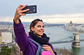 Young woman taking a selfie with skyline behind, including Parliament building and Danube River, Budapest, Hungary, Europe