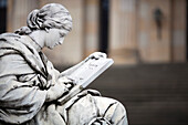 A serene statue of a muse engrossed in reading, located in Gendarmenmarkt, Berlin\'s historic square, surrounded by grand architecture.