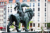 A detailed bronze statue depicting a winged figure riding a lion stands in Gendarmenmarkt, Berlin, near the historic Concert Hall.