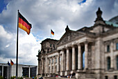 The historic Reichstag building stands prominently in Berlin, adorned with fluttering German flags against a dramatic sky.