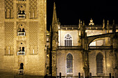 The lighting highlights the intricate details of the Giralda and the nearby Gothic cathedral at night in Seville.