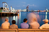 Groups of people enjoy the evening ambiance near the harbor in Barcelona, with waterfront architecture and light reflections.