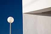 A street lamp stands next to a whitewashed wall, casting a shadow against a bright blue sky in Seville, Spain, showcasing a serene urban moment.