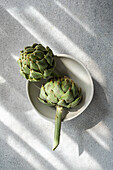 Two fresh artichokes in natural sunlight on a white plate