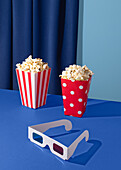 Two popcorn buckets and 3D glasses against a blue background