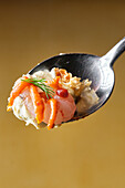 Close-up of a spoon holding a gourmet meal consisting of smoked prawns, mashed potato and a sprig of dill, against a warm golden background