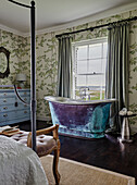 Free-standing bathtub in a bedroom with floral wallpaper and antique furniture