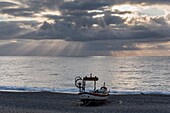 Italien, Ligurien, Noli, Fischerboot am Strand und Lichtstrahlen hinter den Wolken auf dem Mittelmeer