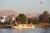 Ägypten,Oberägypten,Niltal,Luxor,große Feluke auf dem grünen linken Nilufer mit den Bergen des Tals der Könige im Hintergrund und einem Heißluftballon am Morgenhimmel