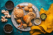 A rustic, teal-colored background with a variety of chai spices and freshly baked muffins, some whole and some broken apart, revealing their fluffy interior. Around the platter are cinnamon sticks, fresh ginger, and small bowls filled with various spices, including black peppercorns, cloves, fennel seeds, and cardamom pods, adding a touch of natural color and texture. A saffron yellow linen napkin adds a pop of warmth and complements the overall rustic aesthetic. The image captures a simple, yet inviting scene, suggesting the warmth of home baking and the comforting aroma of spices.