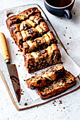 A sliced banana chocolate chip loaf placed on a wooden board beside a knife and a cup of coffee.