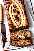 A sliced banana chocolate chip bread with a knife next to it,which has a moist texture and is topped with fresh banana slices