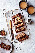 A sliced banana chocolate bread served on a rustic wooden board, alongside two cups of coffee and a small dish of brown sugar.