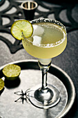 Lime Margarita with salt rim on black surface, halloween setting, lime slices on black tray