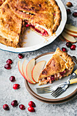 A freshly baked apple and cranberry cake lies in a white cake tin with a slice of cake on a plate next to it. The pie has a golden brown crust and a sweet, juicy filling. The slice of pie is garnished with a few apple slices and a fork lies on the plate. A few cranberries are scattered around the pie and plate, adding a touch of colour to the scene. The picture was taken on a custom rustic grey background