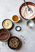 An assortment of cooking ingredients including flour, sesame seeds, flax seeds, sunflower seeds, oil, and salt arranged on a textured surface.