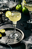 Lime Margarita with salt rim on black surface,Halloween setting,lime slices on black tray
