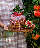 Young girl in checkered dress holding a beautifully decorated cake on a wooden tray,topped with pink icing and fresh pomegranate,rustic mood,outdoor celebration or autumn gathering,natural sunlight pomegranate orchard