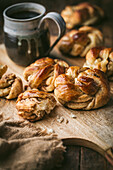 A rustic wooden board is laden with freshly baked cardamom buns, their golden-brown surfaces glistening with a sweet glaze. The rolls are arranged in a tempting array, with some pulled open, revealing the soft, fluffy interior. A rustic ceramic mug, filled with dark coffee, sits beside the rolls. The warm, inviting tones of the image evoke a sense of homeliness and indulgence, focusing on the deliciousness of the baked goods and the pleasure of a simple morning ritual.