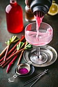 Action shot of pink rhubarb cocktail being poured from shaker into beautiful coupe glass with fresh rhubarb and red syrup in background