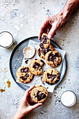 A plate of freshly baked chocolate chip cookies with two hands reaching for them, accompanied by a glass of milk and a small dish of dipping sauce.