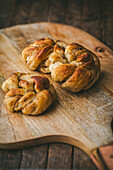 Three freshly baked, golden brown cardamom buns on a wooden chopping board. The background is a dark brown wooden table, giving the image a warm and inviting look that evokes a sense of comfort and indulgence