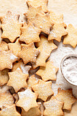 Star-shaped shortcrust cookies dusted with a light sprinkle of powdered sugar. On the right side, a bowl filled with powdered sugar can be seen, used to make these treats even more irresistible. Top view.