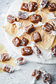 A close-up of homemade caramel sweets on a marble surface. The sweets are wrapped in white parchment paper and sprinkled with sea salt. They are arranged on a piece of brown parchment paper standing on a white marble cake stand. More caramels can be seen in the blurred background, hinting at a delicious treat and emphasising the simplicity and beauty of homemade sweets