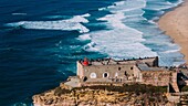 Luftaufnahme von Touristen am historischen Leuchtturm Sao Miguel Arcanjo mit Blick auf die atemberaubende Küste von Nazare mit riesigen Wellen,die eine spektakuläre Aussicht genießen,Nazare,Oeste,Estremadura,Portugal,Europa