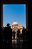 Tourists pass through an archway, revealing Saint Peter's Basilica under a clear sky in Vatican City, showcasing its grand architecture.