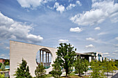 The contemporary German Chancellery building stands in Berlin, surrounded by greenery under a blue sky with fluffy clouds.