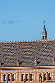 A vibrant skyline view showing La Giralda tower rising behind a historic rooftop in Seville, Spain. Bright blue sky complements the architectural details of Plaza de Espana.