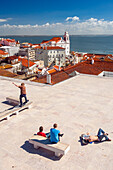 A group of young people enjoy the sun at Santa Luzia viewpoint, overlooking the scenic rooftops and the Tagus river in Lisbon.