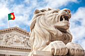 The majestic lion sculpture stands proudly before the neoclassical facade of Sao Bento Palace under a cloudy sky in Lisbon.