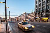 Cars navigate the busy streets of Praca dos Restauradores in Lisbon as twilight sets over the vibrant cityscape.