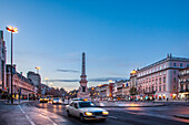 Cars navigate the busy streets of Praca dos Restauradores in Lisbon as twilight sets over the vibrant cityscape.